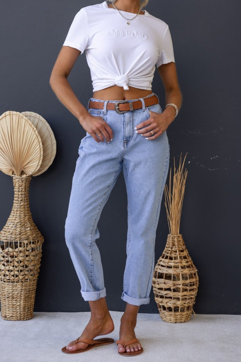 Stylish woman wearing Dori Mom Jeans with rolled cuffs and a knotted white t-shirt, posing against a dark wall.