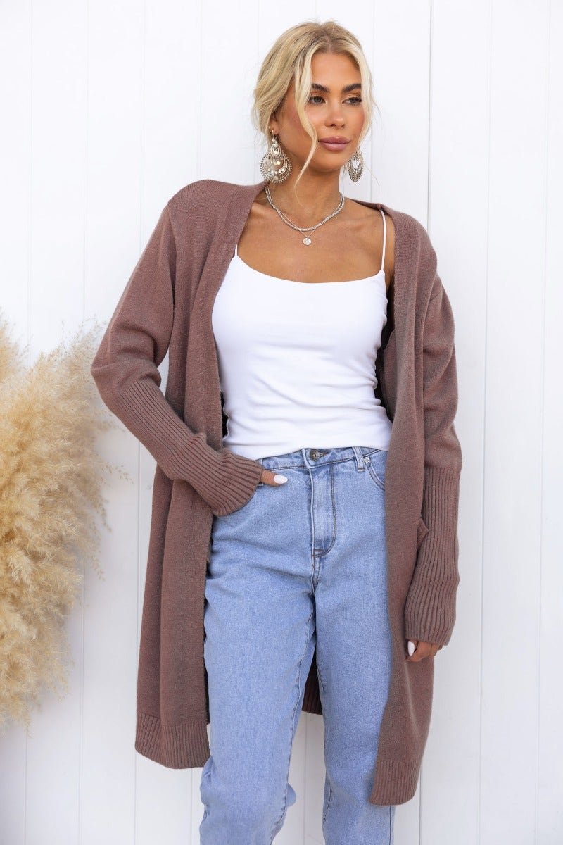 Model wearing a longline ribbed knit Motherly Cardigan in brown, styled with a white tank top and light blue jeans.