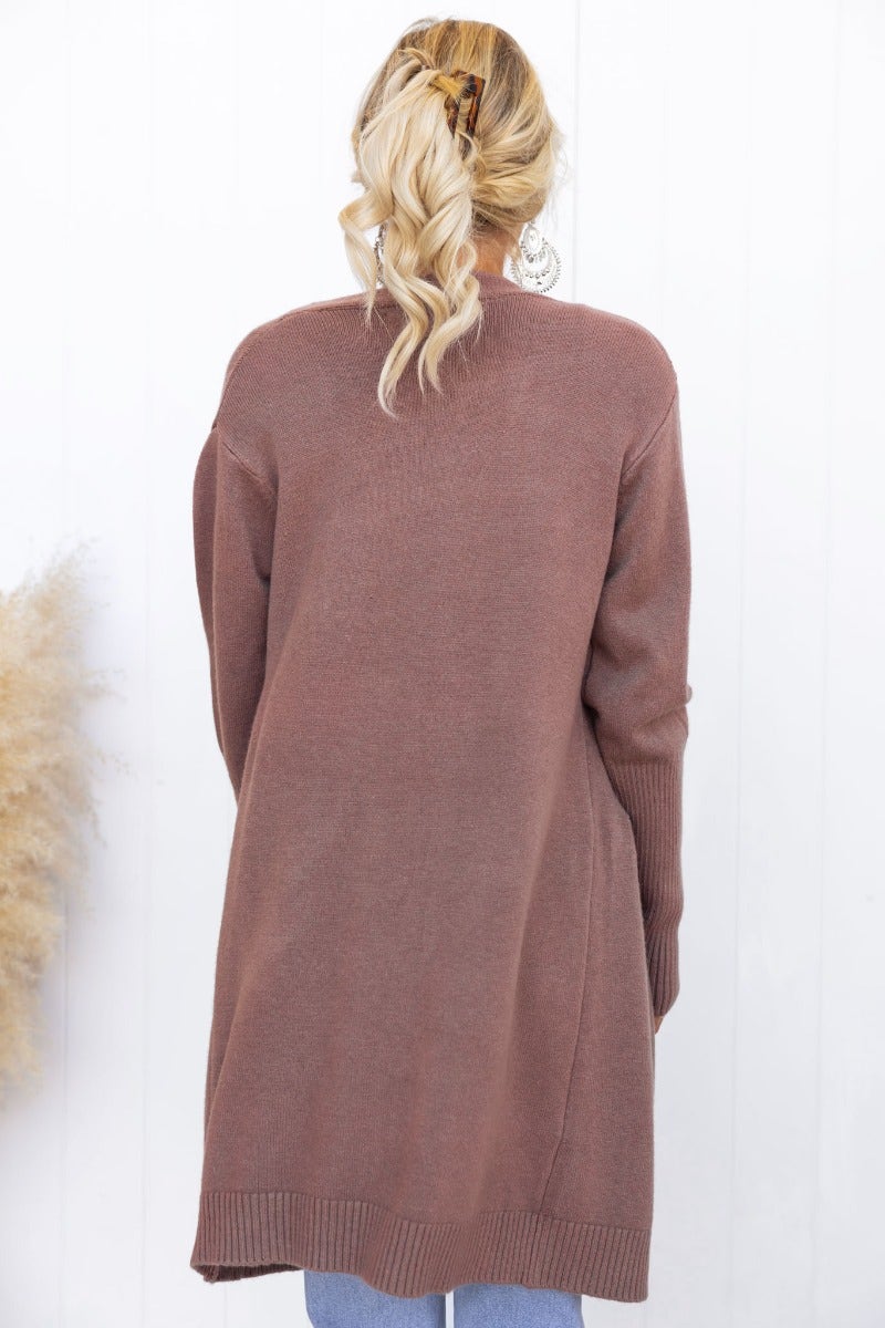 Back view of a woman wearing a longline pink Motherly Cardigan with ribbed knit and pockets.