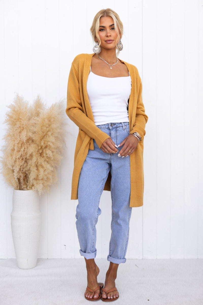 Model wearing a mustard Motherly Cardigan over a white tank top, paired with light blue jeans and sandals.
