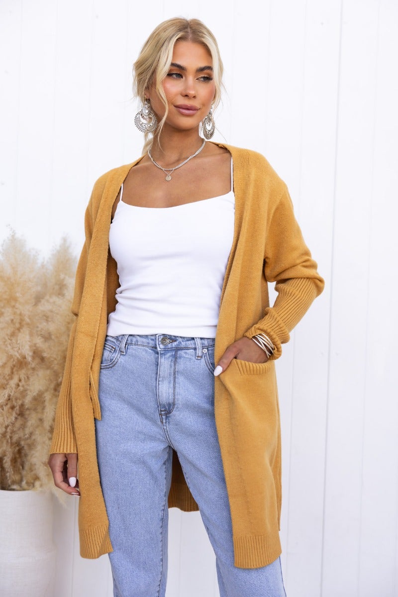 Stylish woman in a mustard Motherly Cardigan, paired with a white top and jeans, showcasing its longline fit and front pockets.
