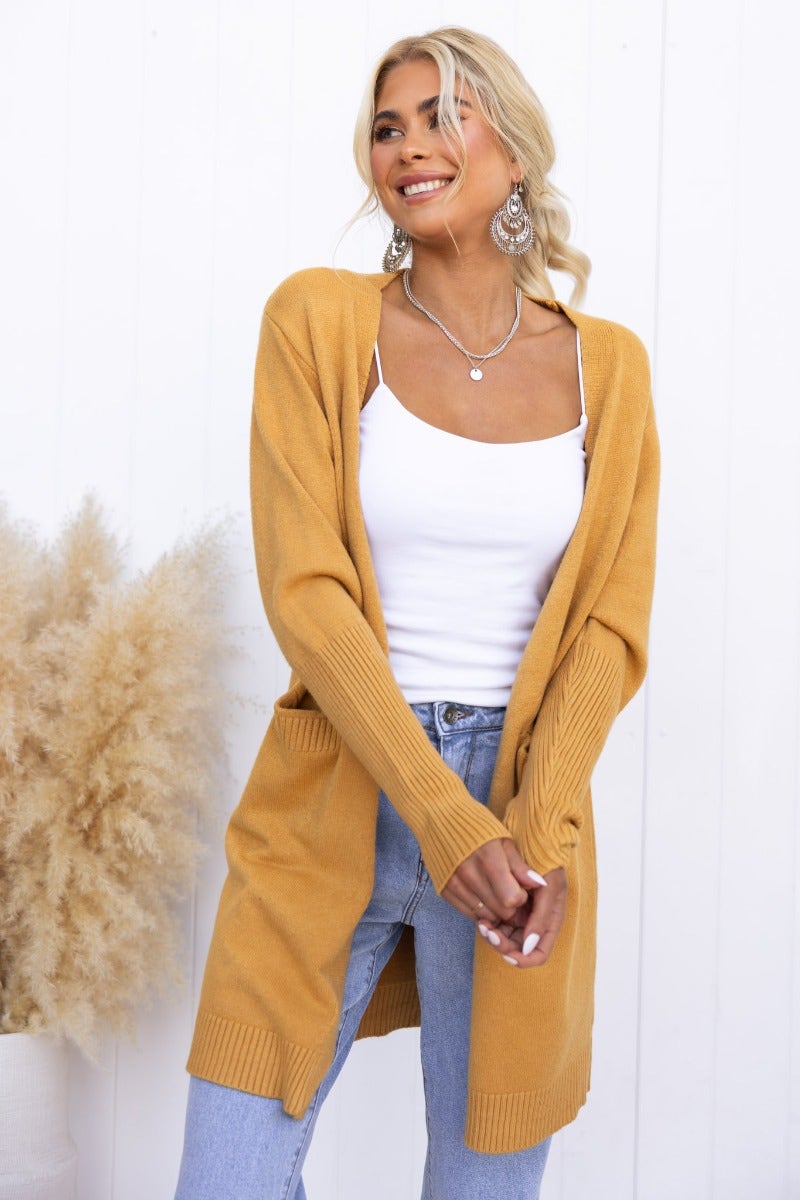 Smiling woman wearing a mustard Motherly Cardigan over a white tank top, styled with light denim and statement earrings.