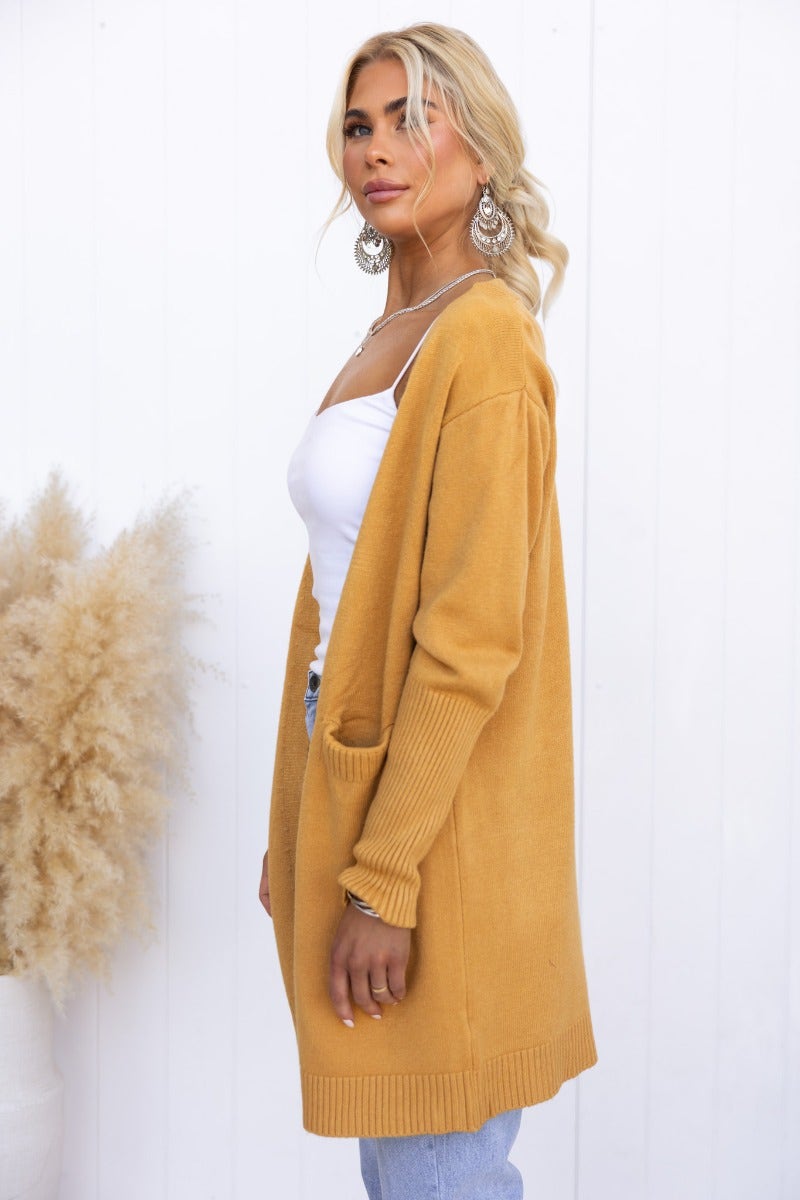 Stylish woman in a mustard Motherly Cardigan with front pockets and a longline fit, paired with a white top and jeans.