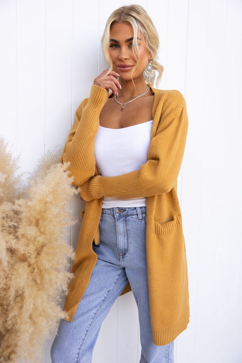 Woman in a mustard longline ribbed cardigan with pockets, styled with a white top and blue jeans.