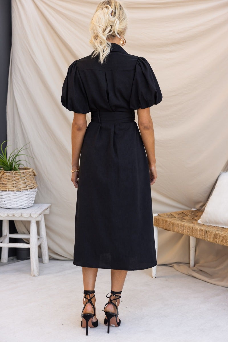 Back view of a woman wearing a black midi dress with puff sleeves and a removable fabric belt.