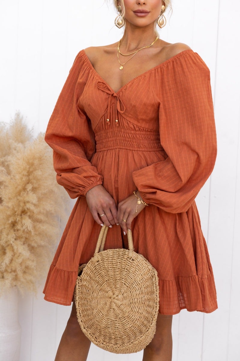 Model wearing an off-shoulder rust-colored cotton dress with tie detail and puff sleeves, holding a woven bag.
