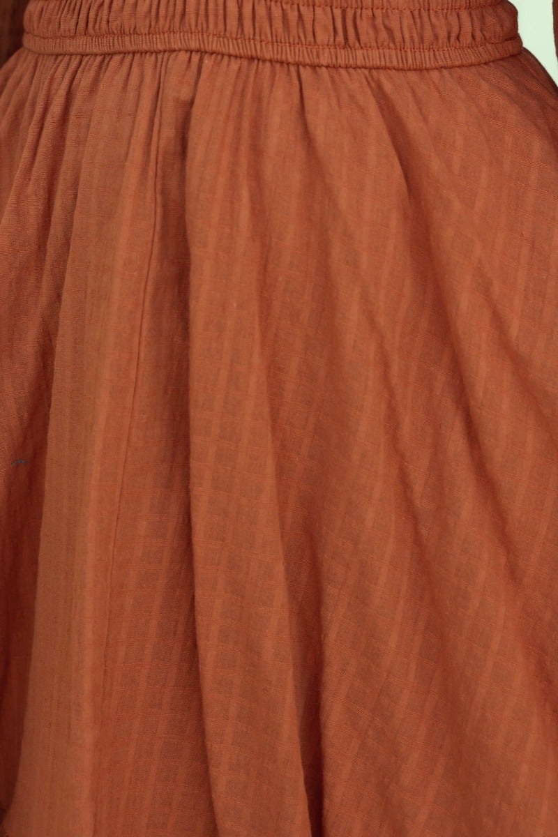 Close-up of rust-colored fabric detail with elastic waist, featuring soft textured pleats in a flowing design.