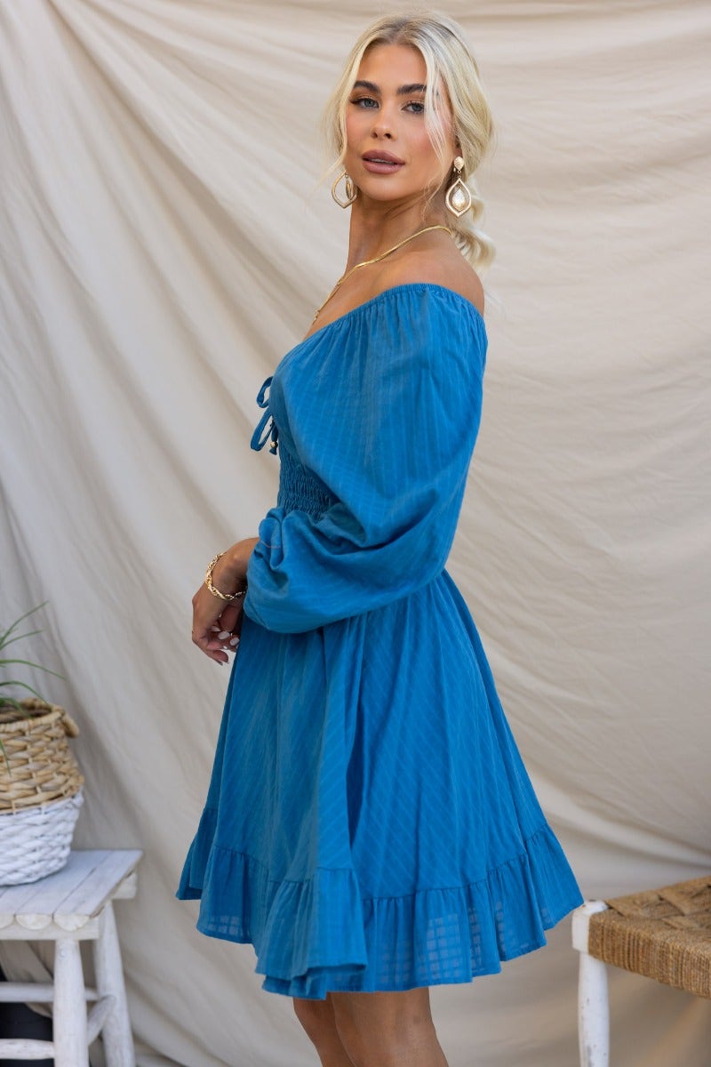 Blue cotton getaway dress featuring elastic sleeves, tie neckline, and ruffled hemline, styled in a trendy off-shoulder look.