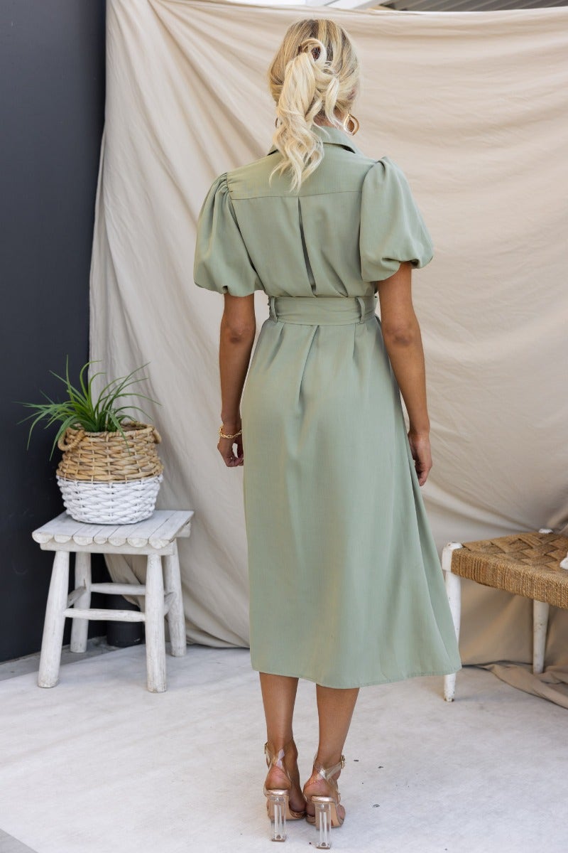 Back view of Sorrento Midi Dress in sage green with puff sleeves and removable belt, styled in a modern setting.