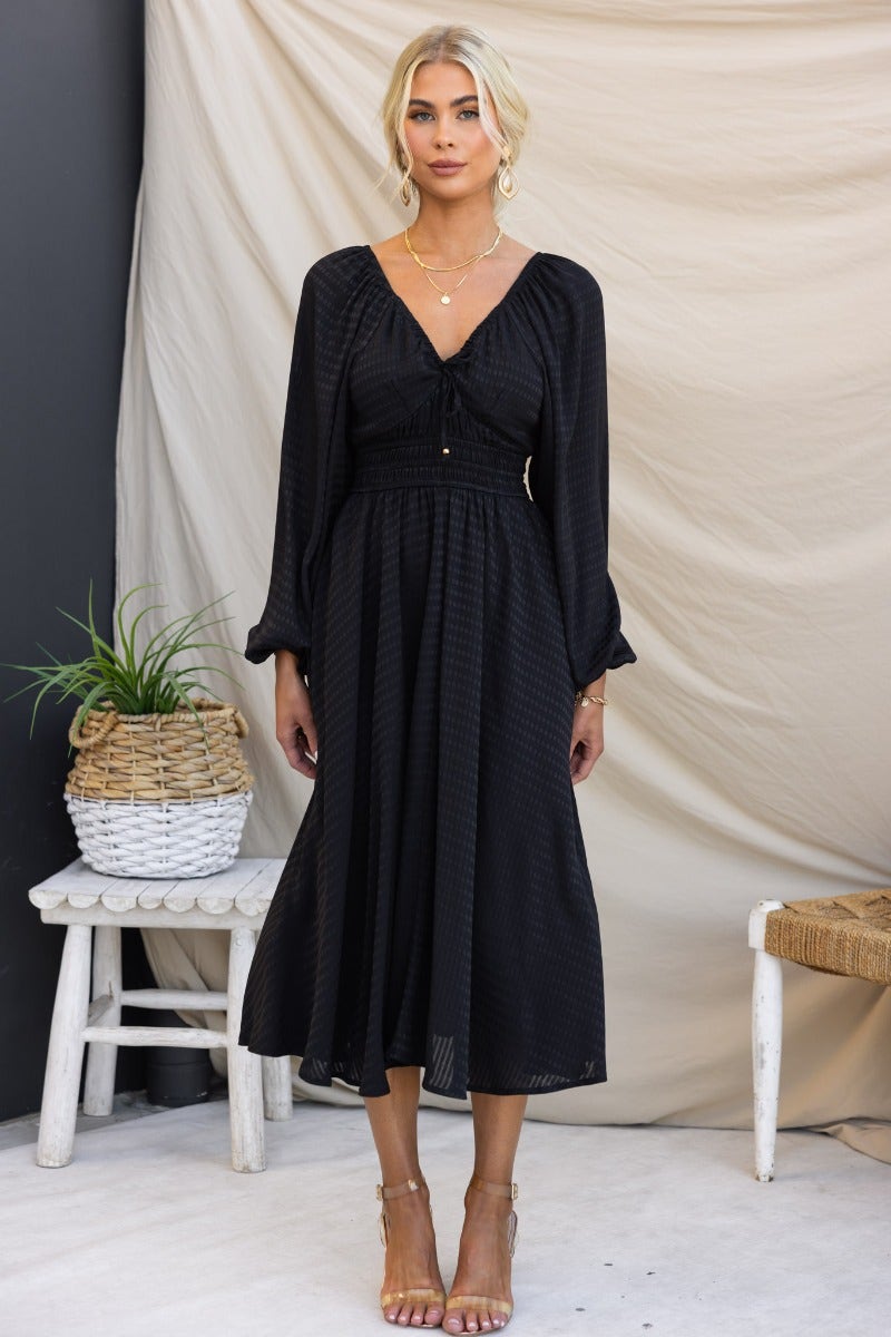 Black textured midi dress with elastic sleeves and tie neckline detail, styled with pockets and a flattering fit.