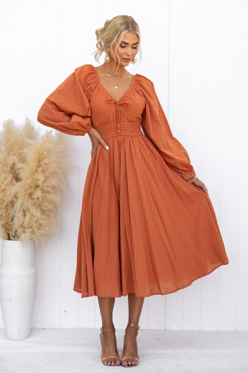 Hemingway Midi Dress in burnt orange featuring puff sleeves, a tie neckline, and a flowy skirt, styled with chic accessories.