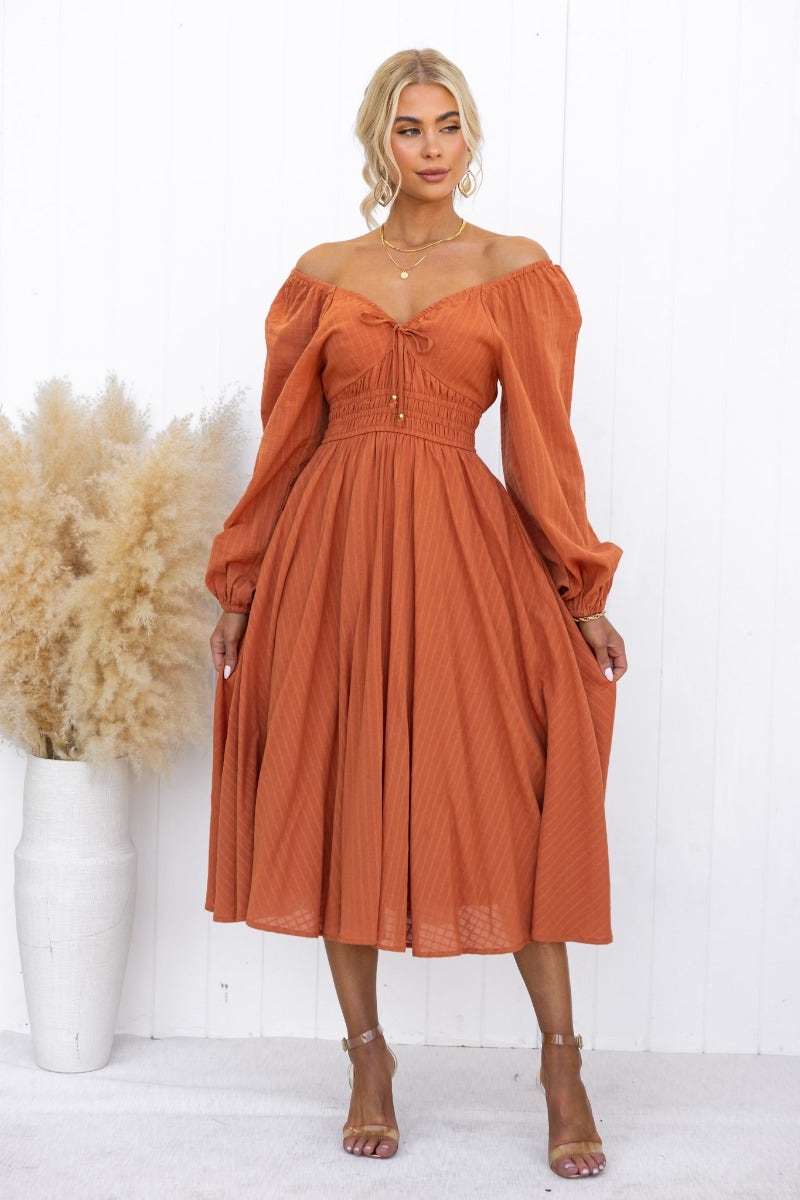 Model wearing an orange Hemingway midi dress with off-shoulder sleeves and tie detail, standing next to a potted plant.