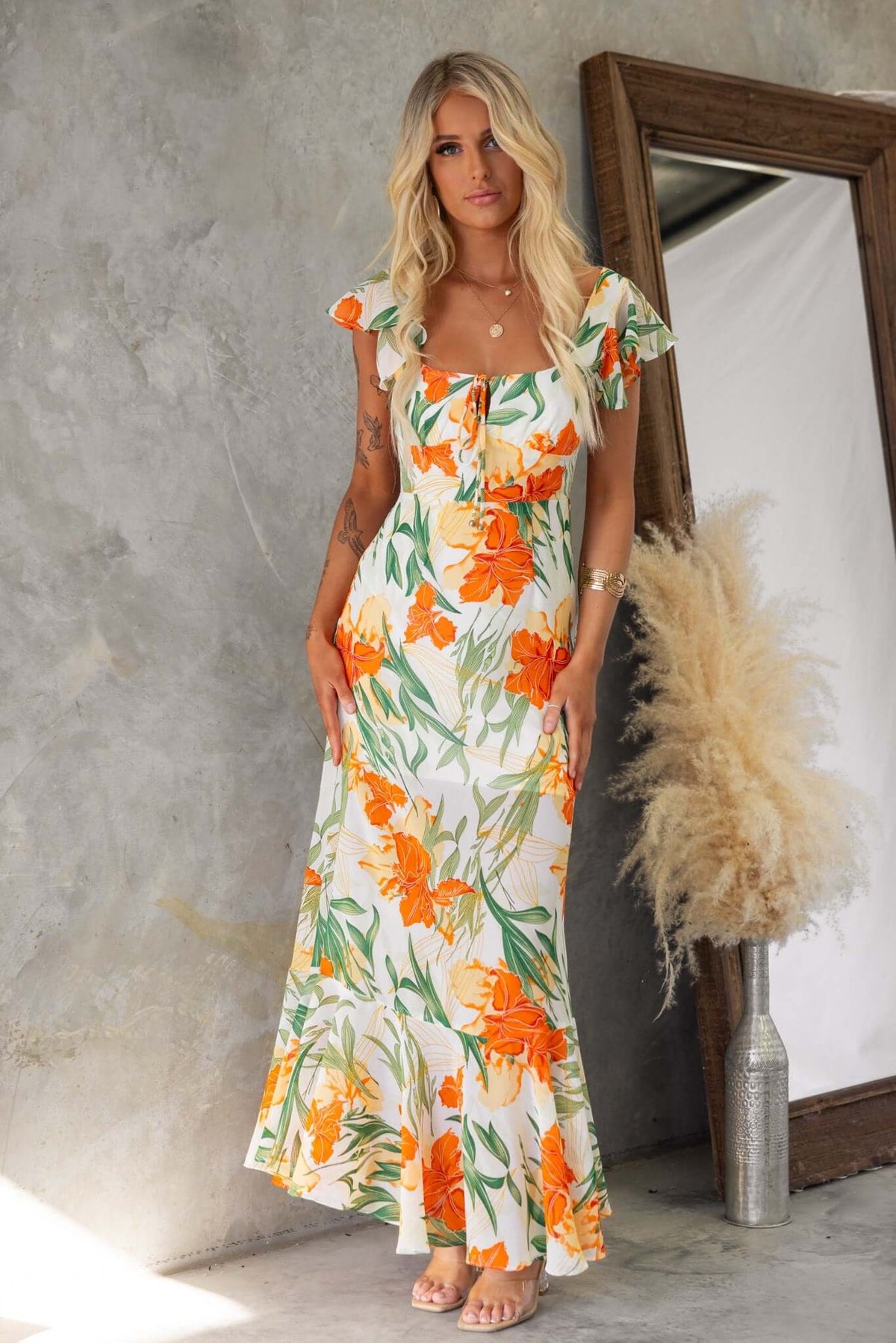 Floral print Tranquil Maxi Dress with V-neckline and tie detail, featuring a model standing against a textured backdrop.