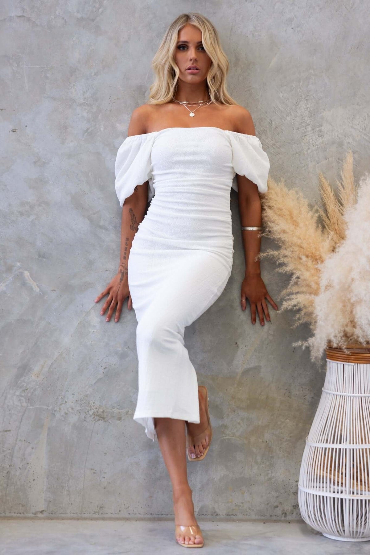 Elegant Topeka Midi Dress in white with off-shoulder design, ruching, and split at back, styled with natural decor.