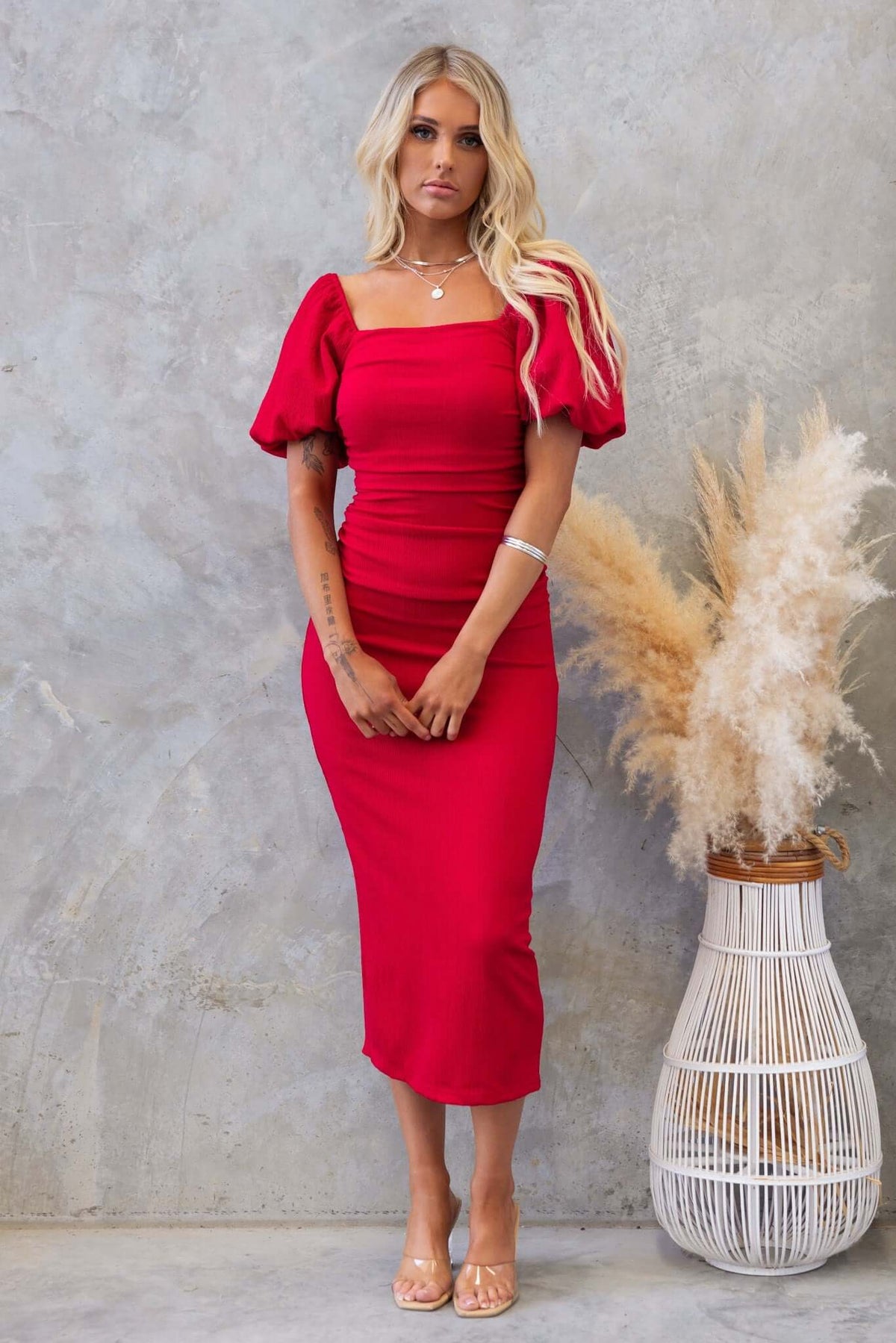 Model wearing a red Topeka Midi Dress with puff sleeves and back split, styled against a textured background.