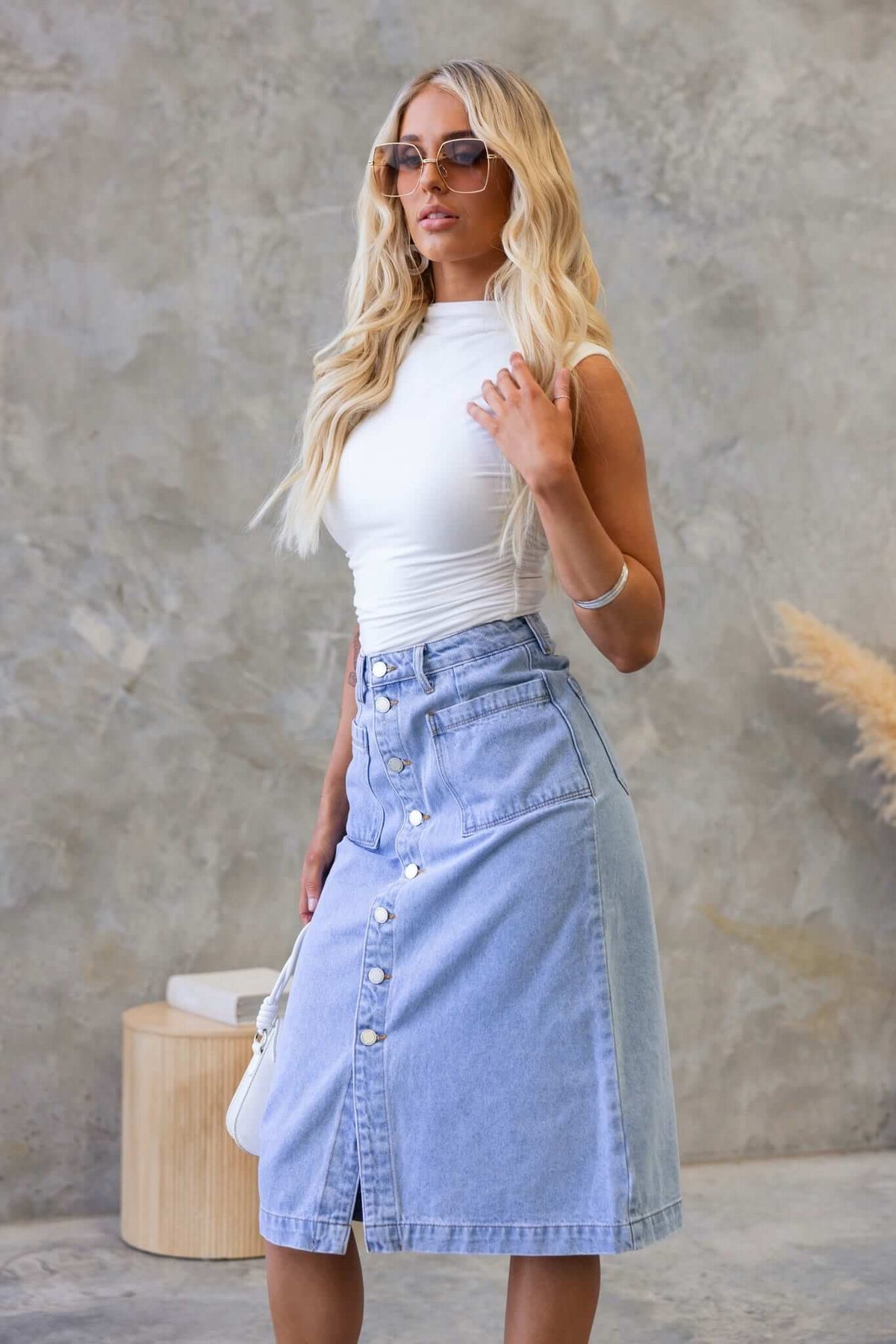 Woman in stylish Soma denim midi skirt with button-up front, paired with a white top and sunglasses, standing against a neutral background.