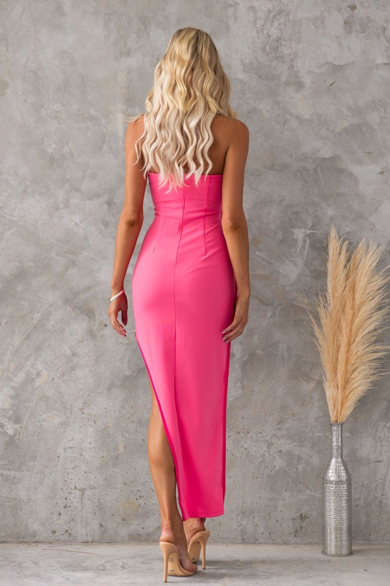 Woman in a strapless pink bodycon maxi dress with an asymmetric hemline, styled with flowing hair and footwear.