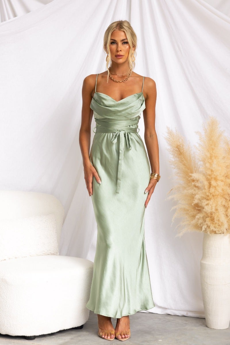 Model wearing a satin green maxi dress with a cowl neckline and adjustable straps, styled with a tie detail at the waist.