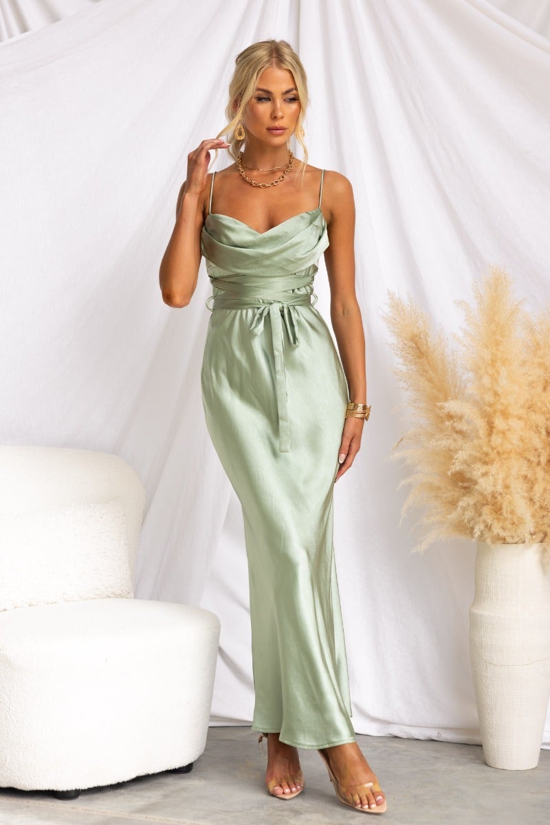 Model wearing a satin cowl neckline maxi dress in sage green with adjustable straps and waist tie detail.