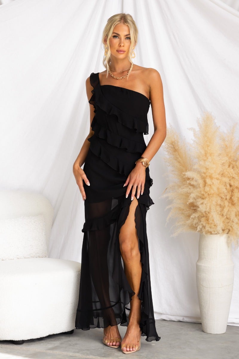 Model wearing a black one-shoulder maxi dress with ruffles and a front split, styled with neutral accessories.