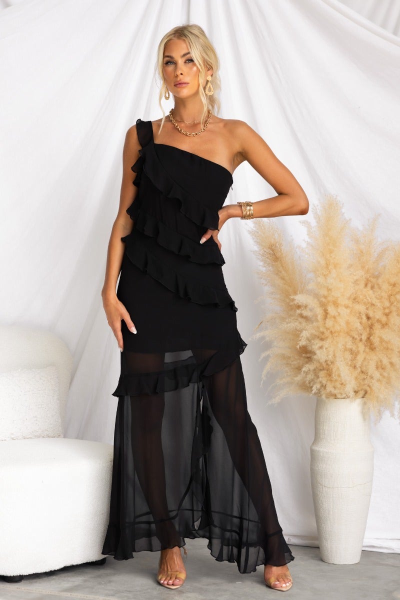 Model wearing a black one-shoulder maxi dress with ruffle detail and side split, styled against a neutral backdrop.