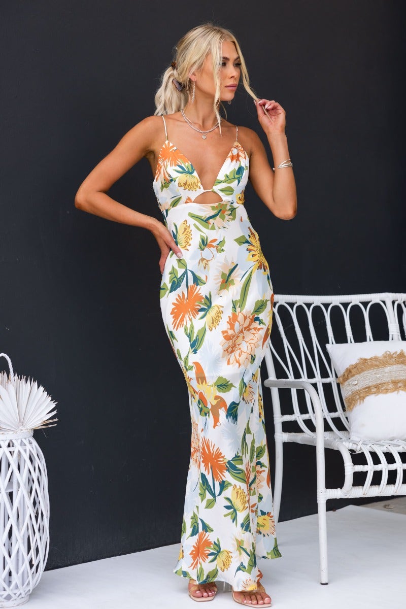 Model wearing a floral satin Disappear Maxi Dress with cut-outs and adjustable straps, styled against a black wall.