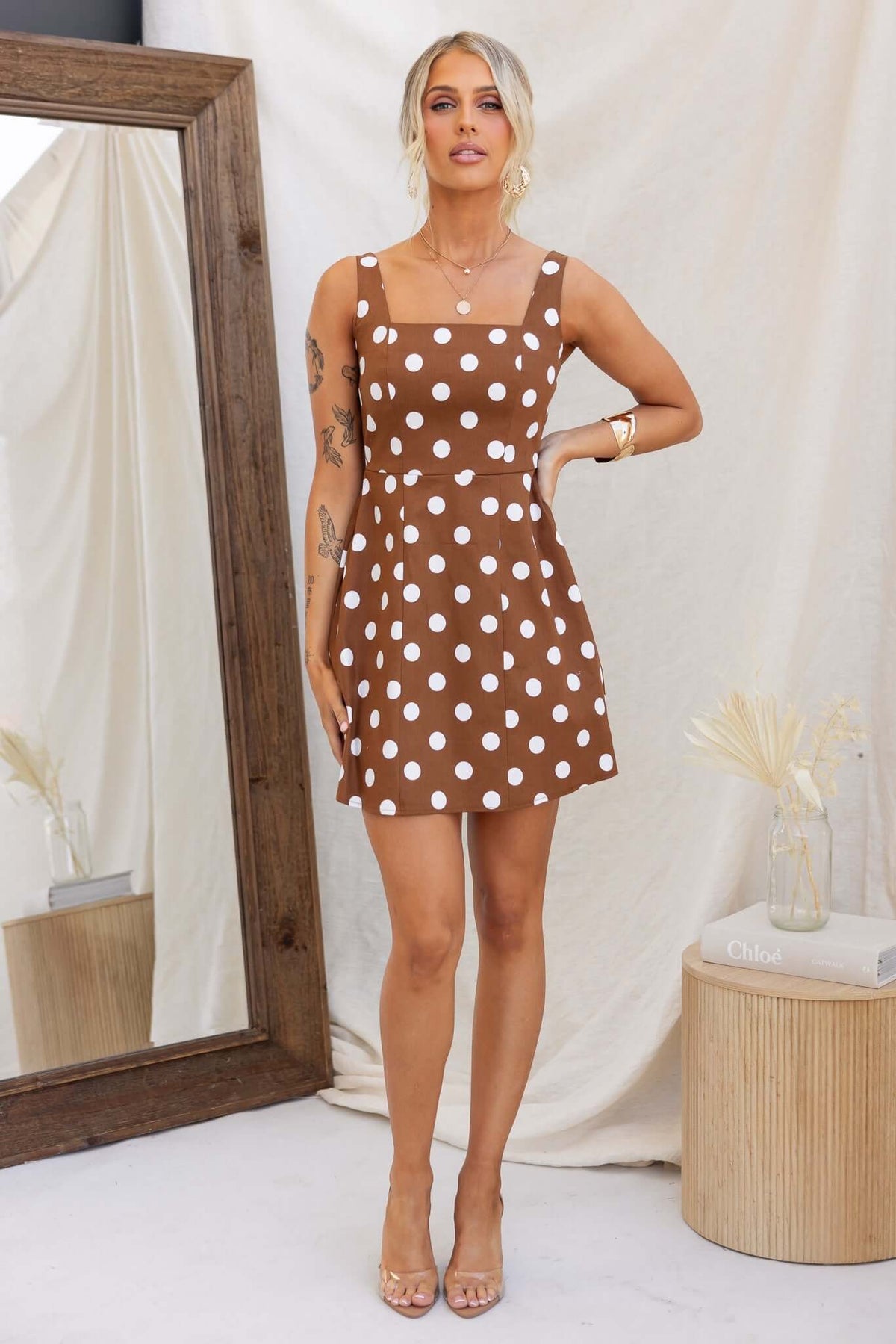 Model wearing Isibel Dress, a brown polka dot dress with adjustable straps and pockets, styled elegantly with a mirror backdrop.
