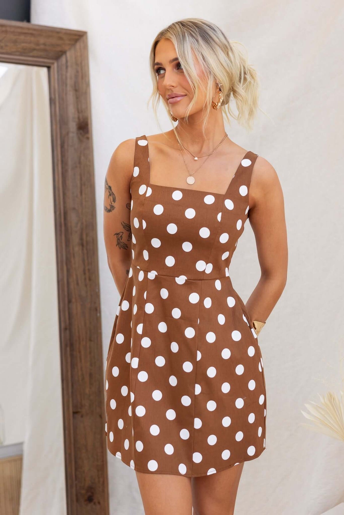 Model wearing Isibel Dress, a brown polka dot dress with adjustable straps and pockets, showcased in a styled setting.