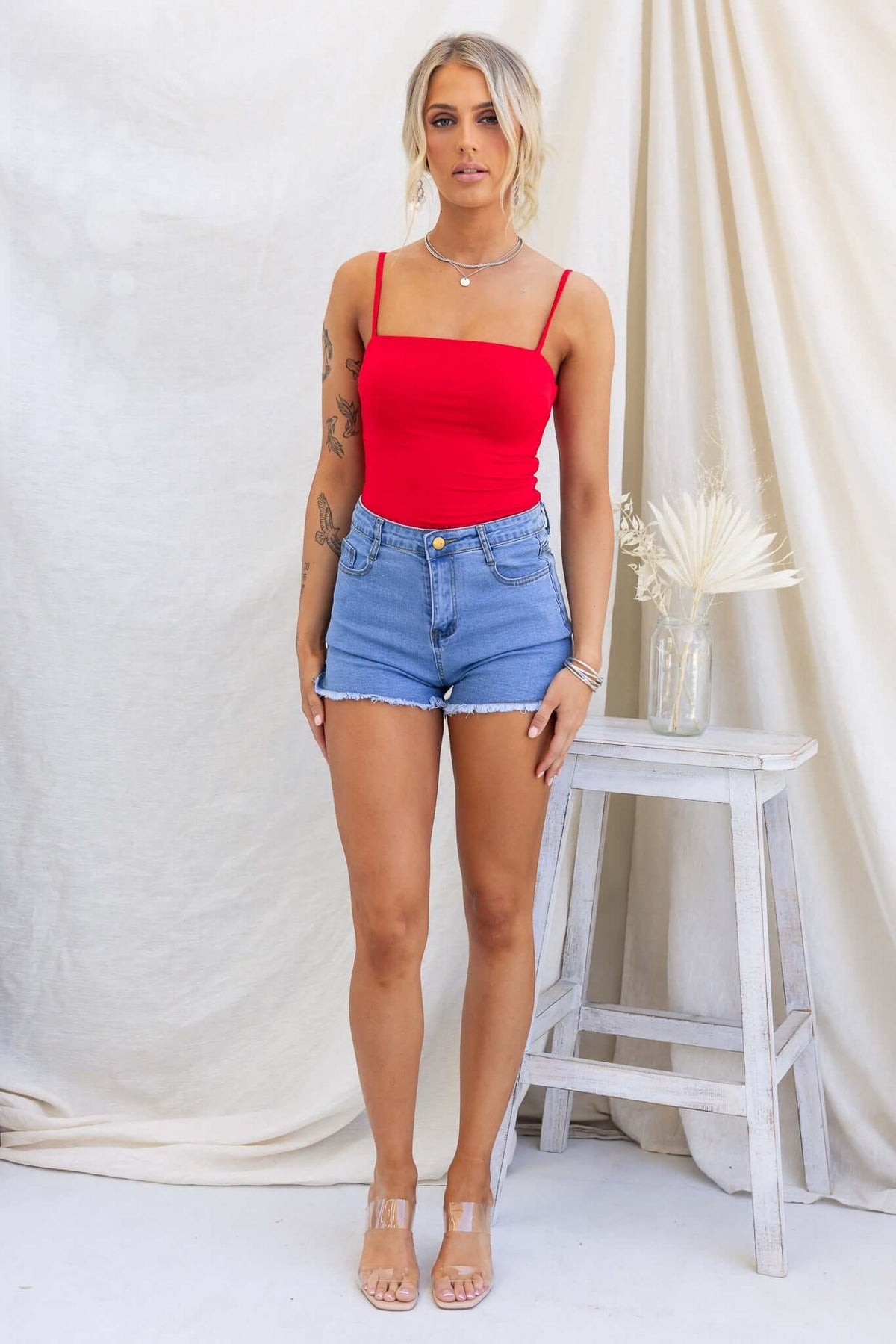 Model wearing a red Brea bodysuit with adjustable straps and denim shorts, showcasing a stylish summer outfit.