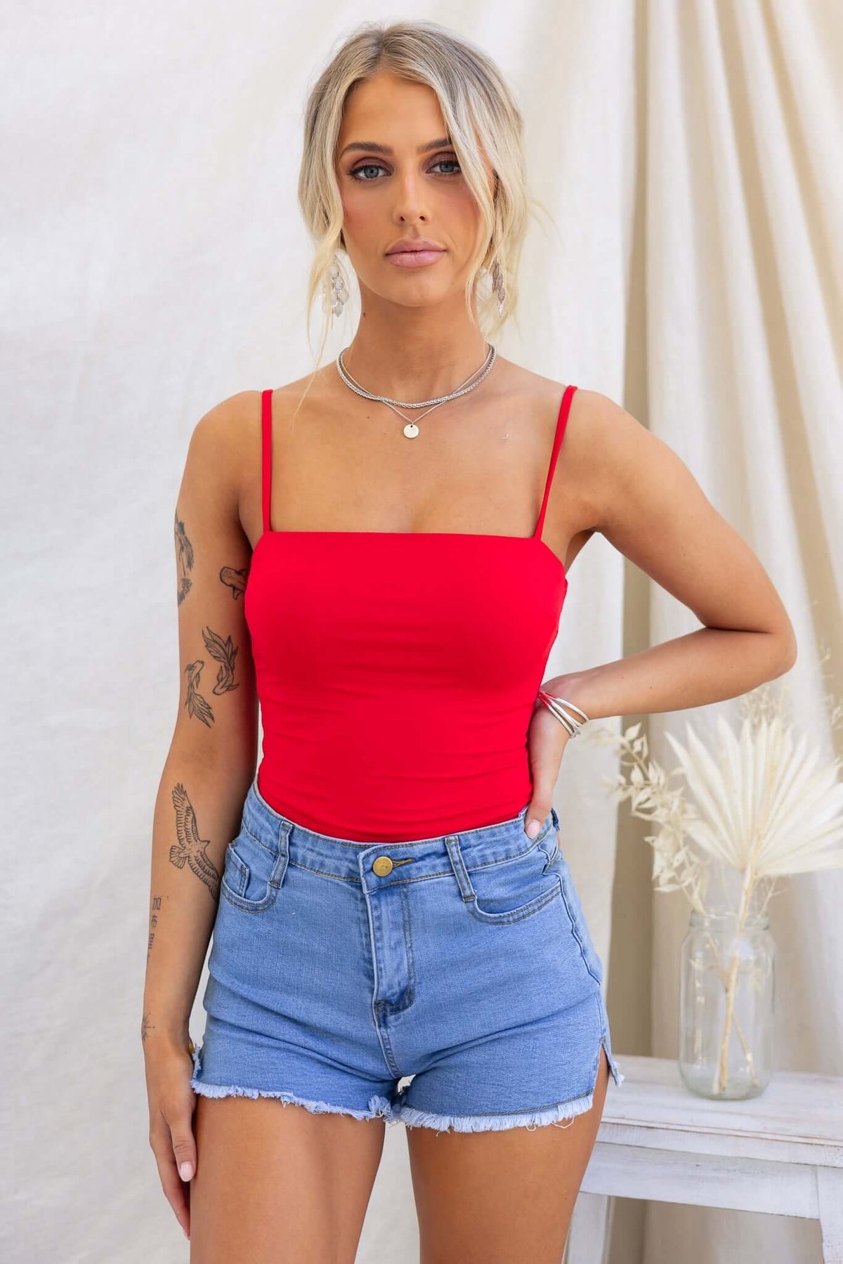 Model wearing a red Brea bodysuit with adjustable straps, paired with denim shorts.