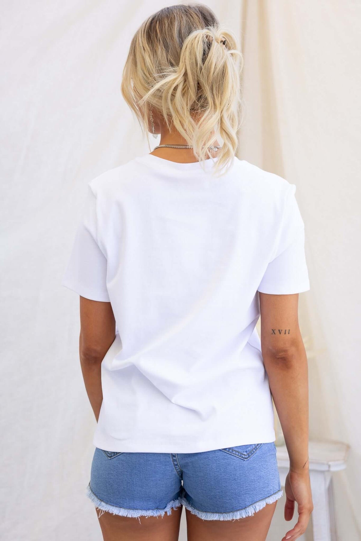 Back view of a woman in a white Doha Tee, paired with denim shorts, showcasing round neckline and casual style.