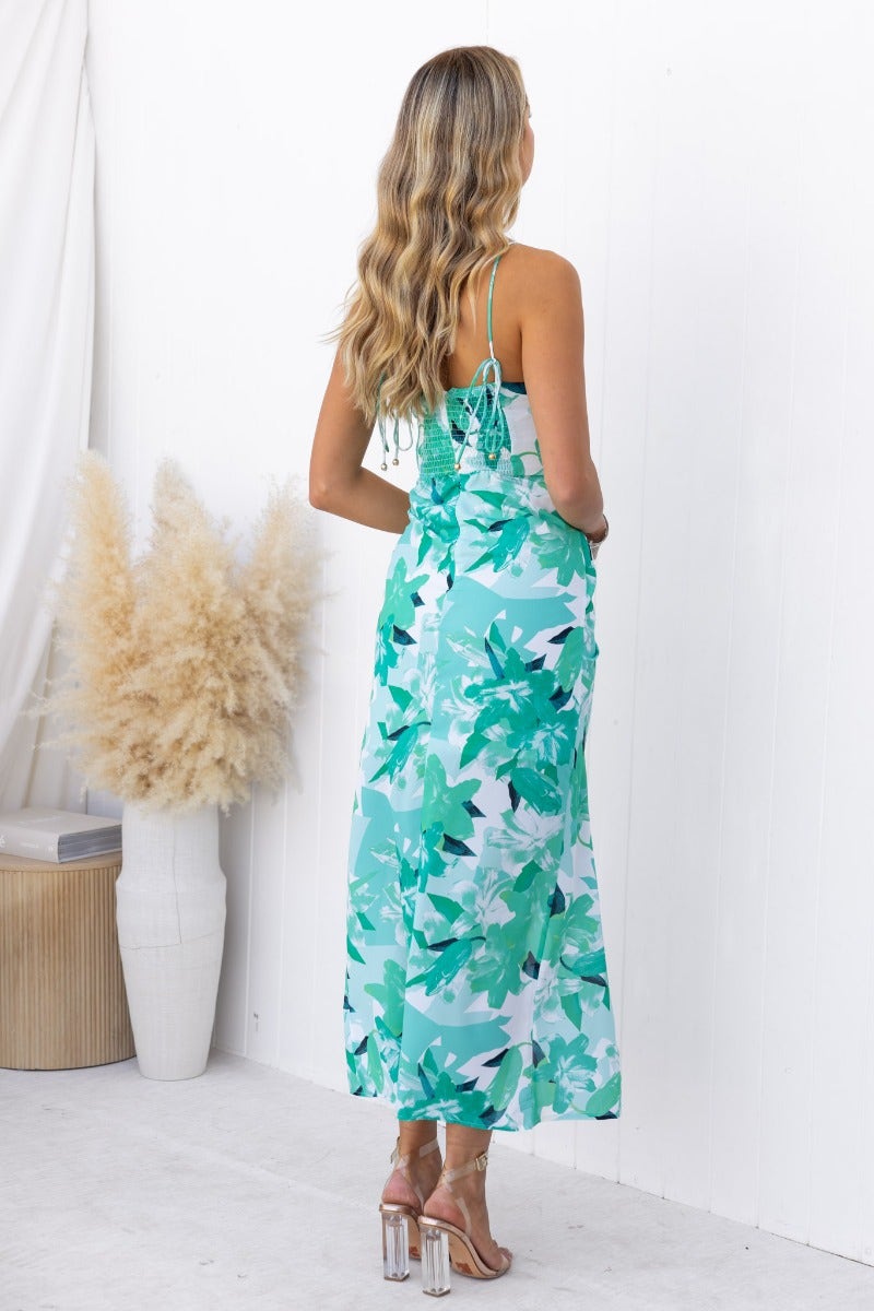 Back view of a floral print vacation ready maxi dress in teal and green, featuring adjustable tie straps and an asymmetrical hemline.