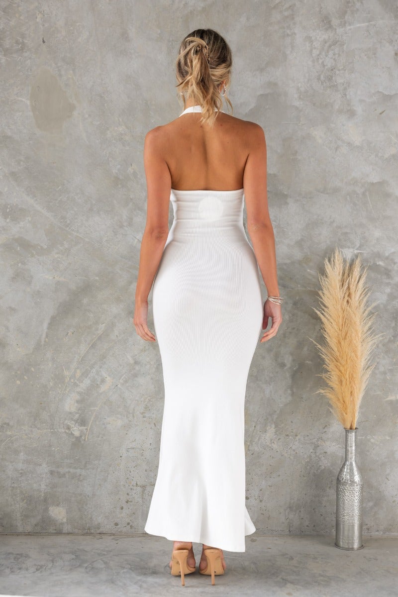 Back view of a woman in a white ribbed halter maxi dress standing against a textured wall, showcasing its elegant silhouette.