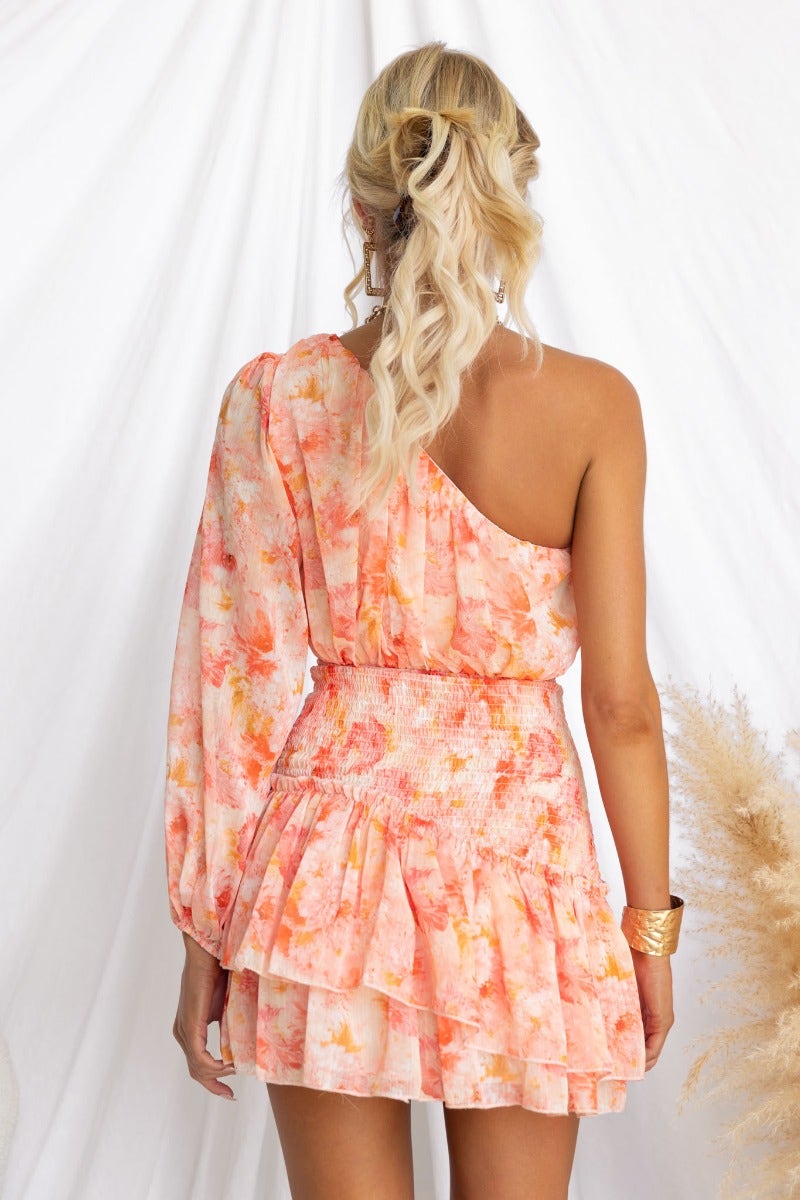 One-shoulder floral print dress in soft peach tones with smocked waist and elastic sleeve cuff.