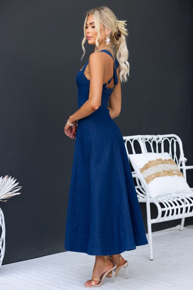 Model wearing a dark navy denim midi dress with adjustable straps and a cross-over back, set against a minimalist background.