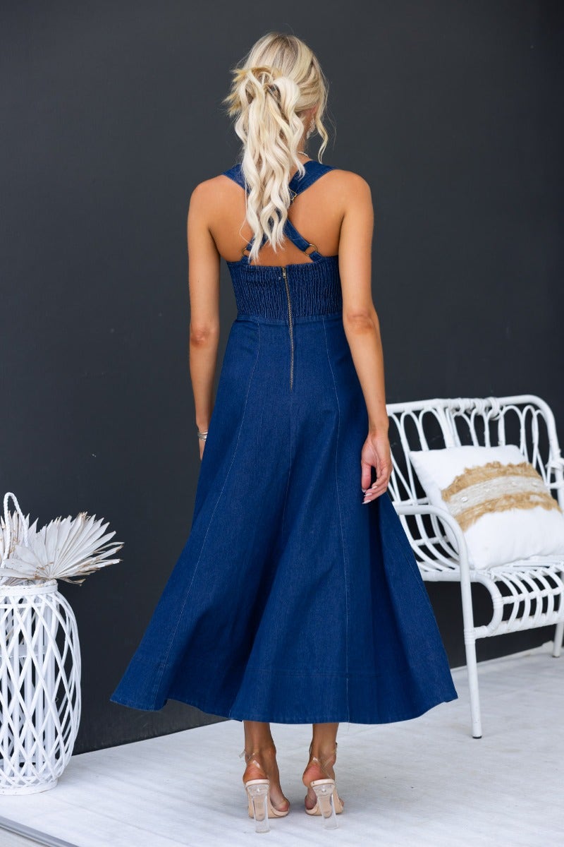 Back view of a woman in a dark navy denim midi dress with adjustable straps and elastic back.