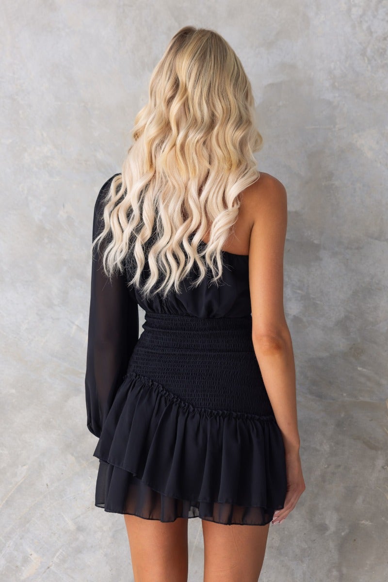 Back view of the Be The Light mini dress featuring a one-shoulder design and ruffled hem, styled with wavy hair.