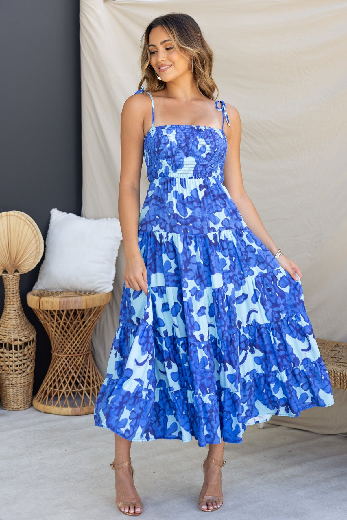 Inessa Midi Dress featuring adjustable tie straps and a vibrant blue floral print, modeled by a woman in a stylish setting.