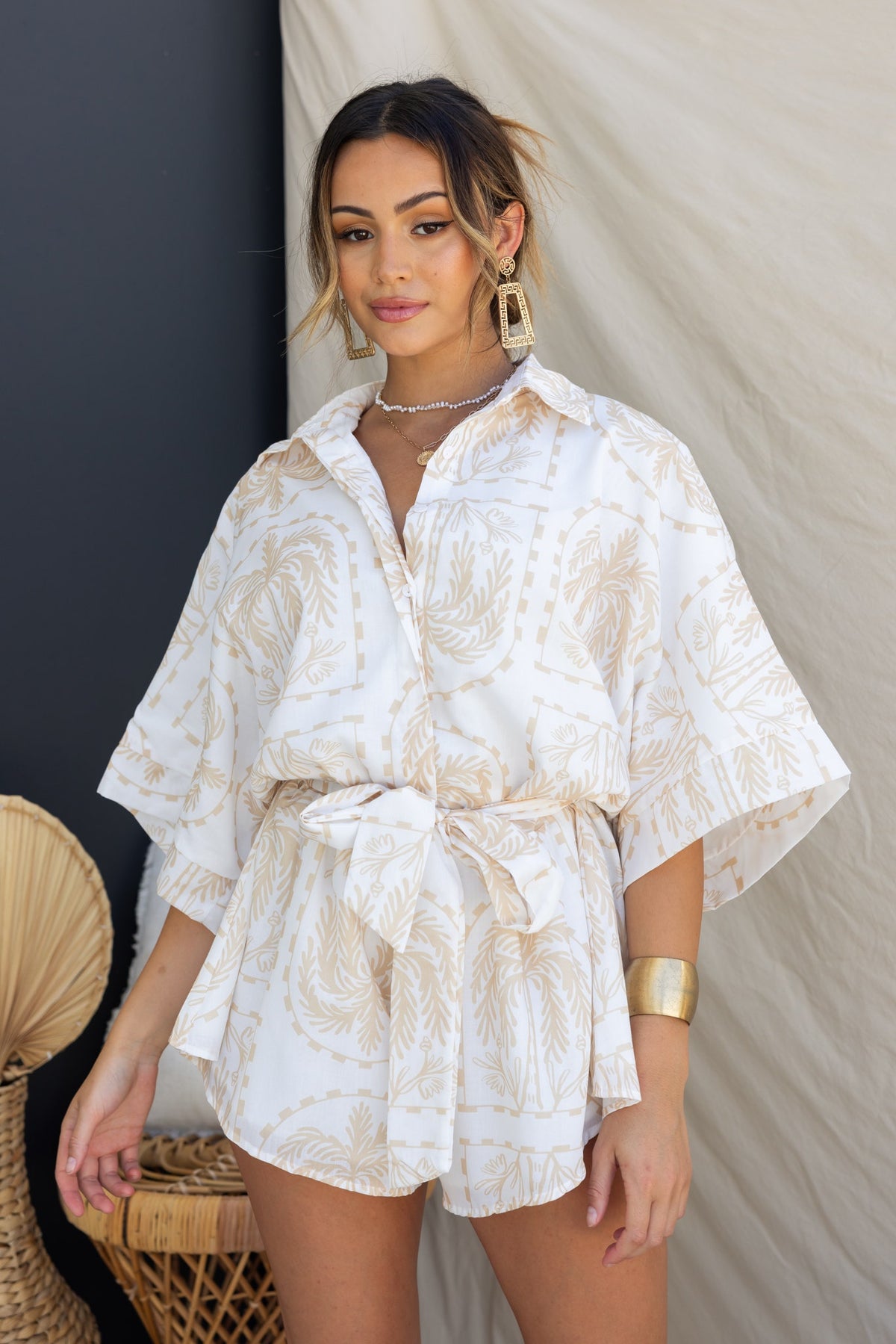 Model wearing white Alli playsuit with collar, button-up front, and fabric tie, styled with oversized fit and gold accessories.