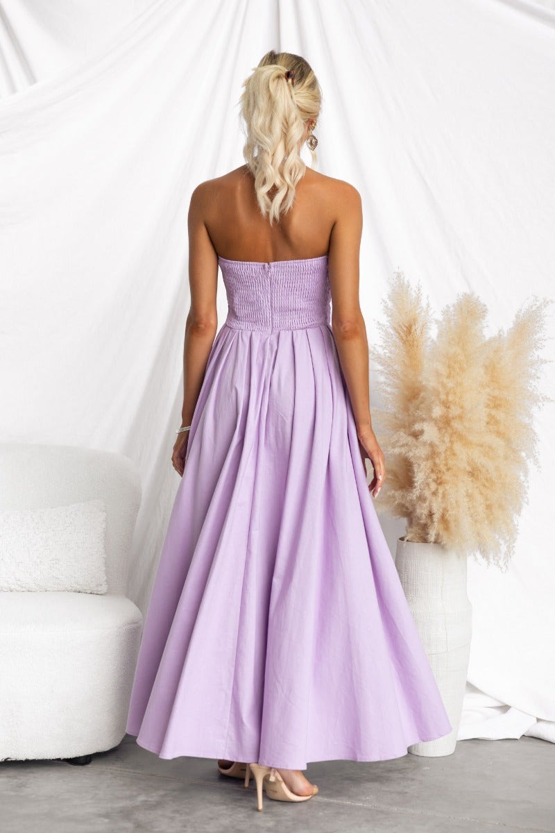 Back view of a woman in a strapless lilac maxi dress, featuring boning and an asymmetric hem.