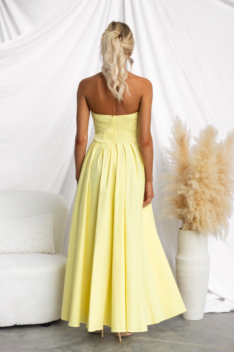 Back view of a woman in a yellow strapless maxi dress, featuring an asymmetric hem and elastic back.