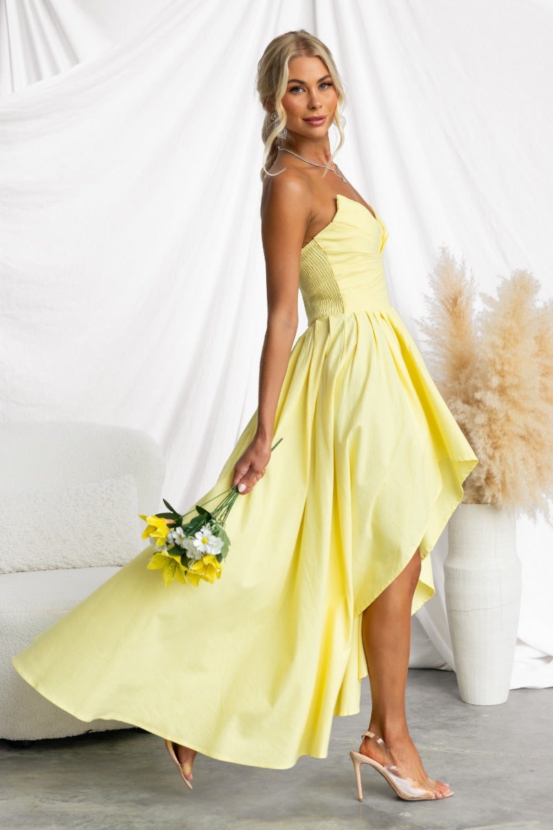 Model wearing a strapless yellow Kiss And Tell Maxi Dress with an asymmetric hem, holding flowers.