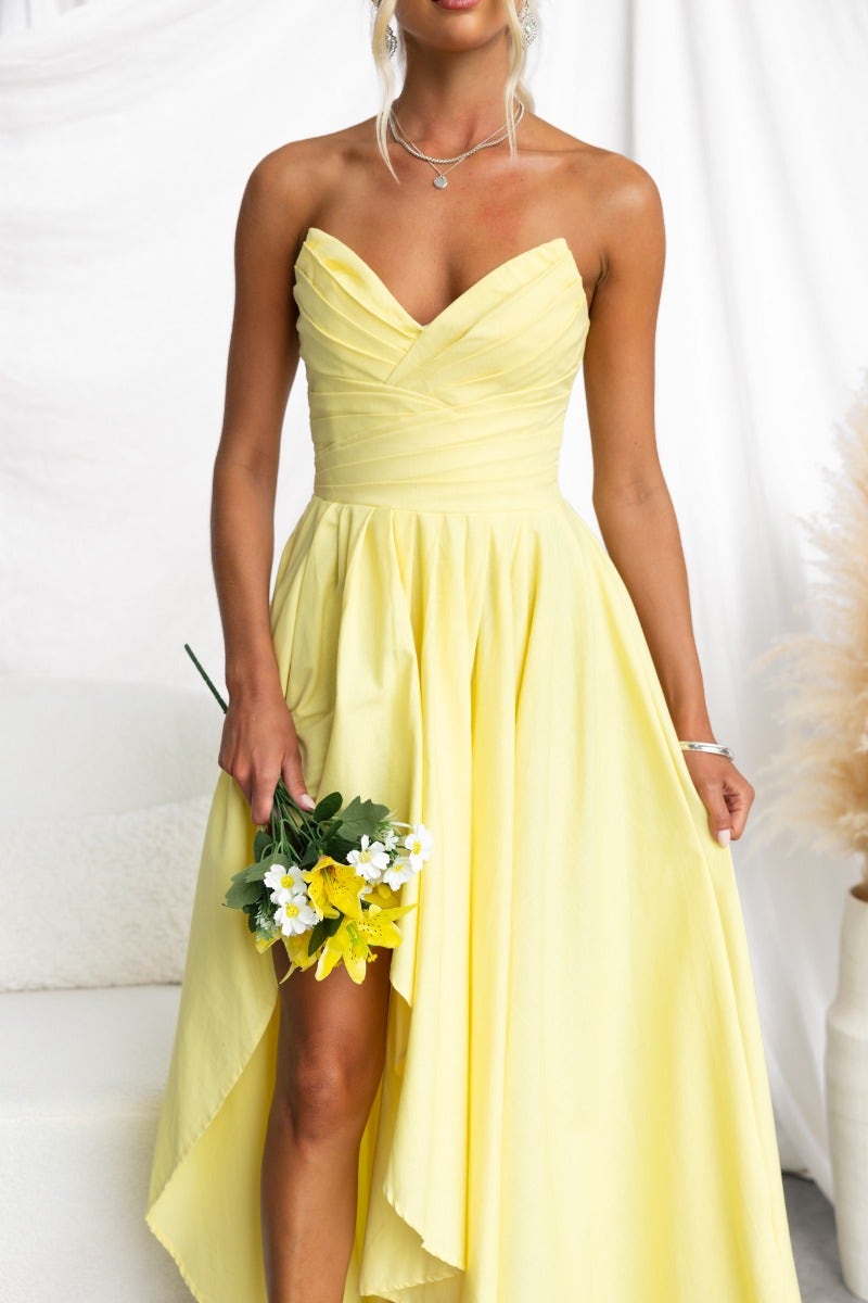 Model wearing a strapless yellow Kiss And Tell maxi dress with an asymmetric hem, holding a bouquet of flowers.