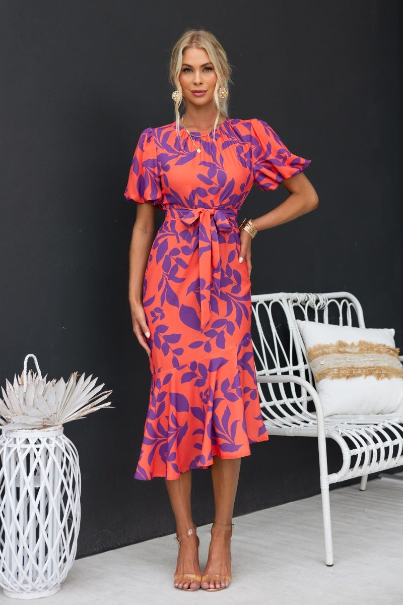 Model wearing Linus Midi Dress, a floral print, orange and purple dress with elastic sleeves and a removable tie.