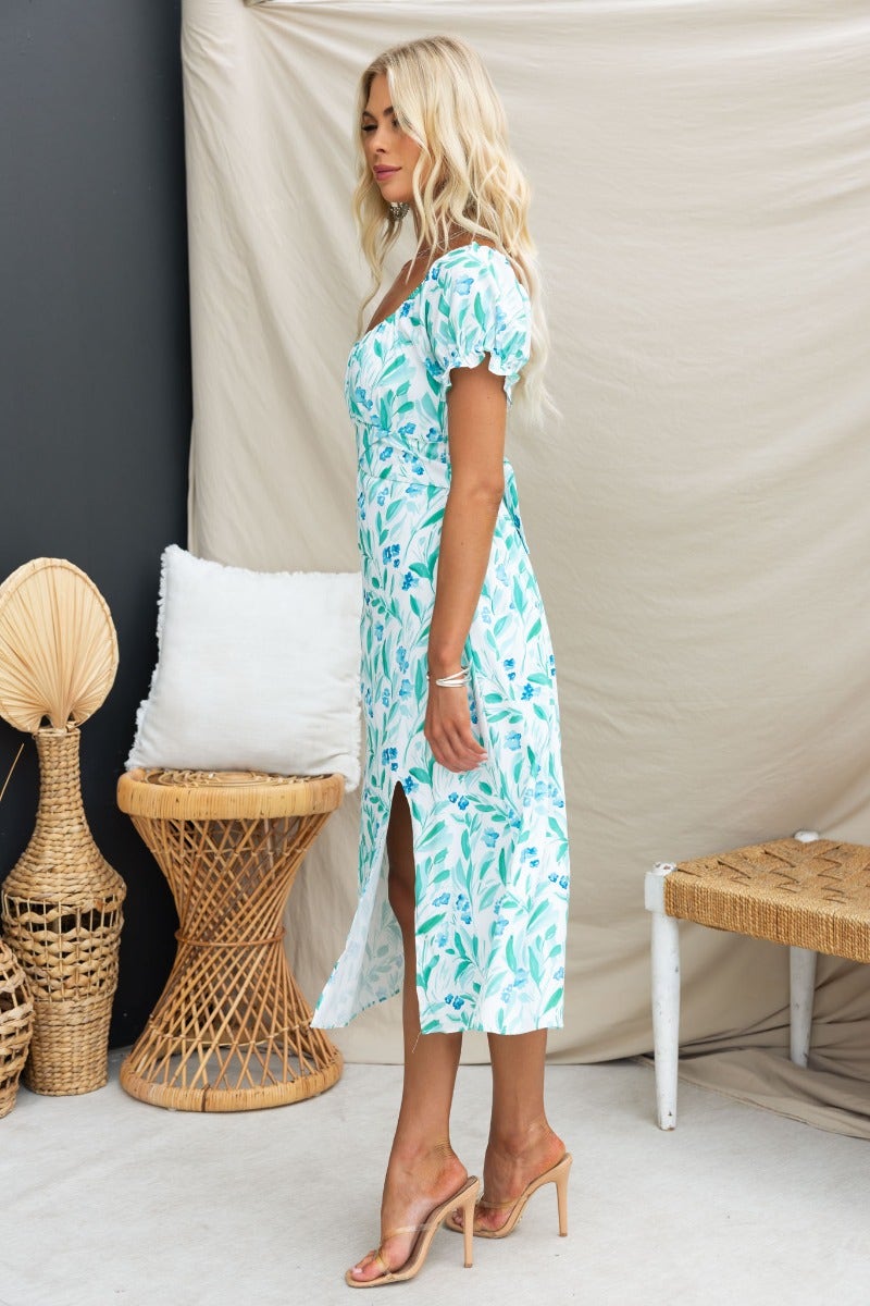 Profile view of a woman in a floral print Teava midi dress with a sweetheart neckline and split, styled in a modern interior.