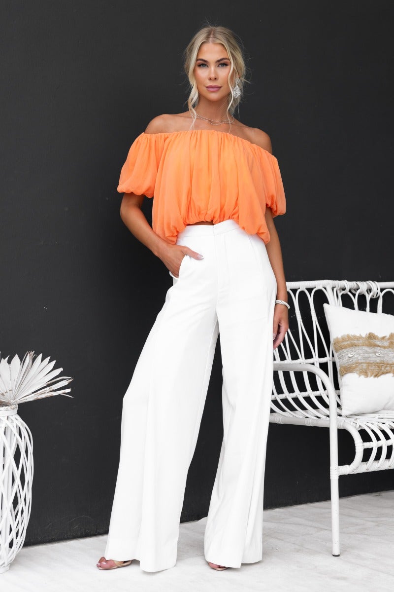 Model showcasing Adanna Top in vibrant orange, off-shoulder design paired with stylish white wide-leg pants.