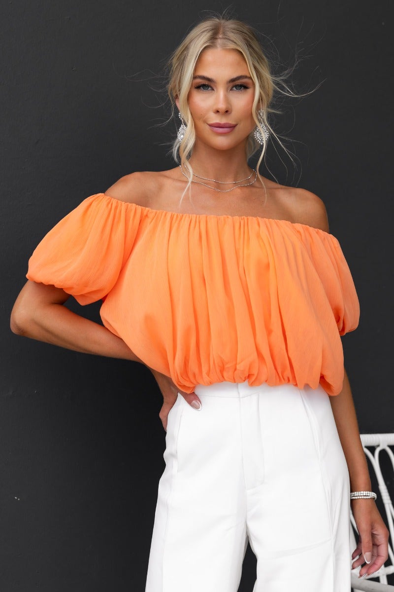 Model wearing an off-shoulder orange chiffon Adanna Top with elastic neckline and sleeves, styled with white pants.