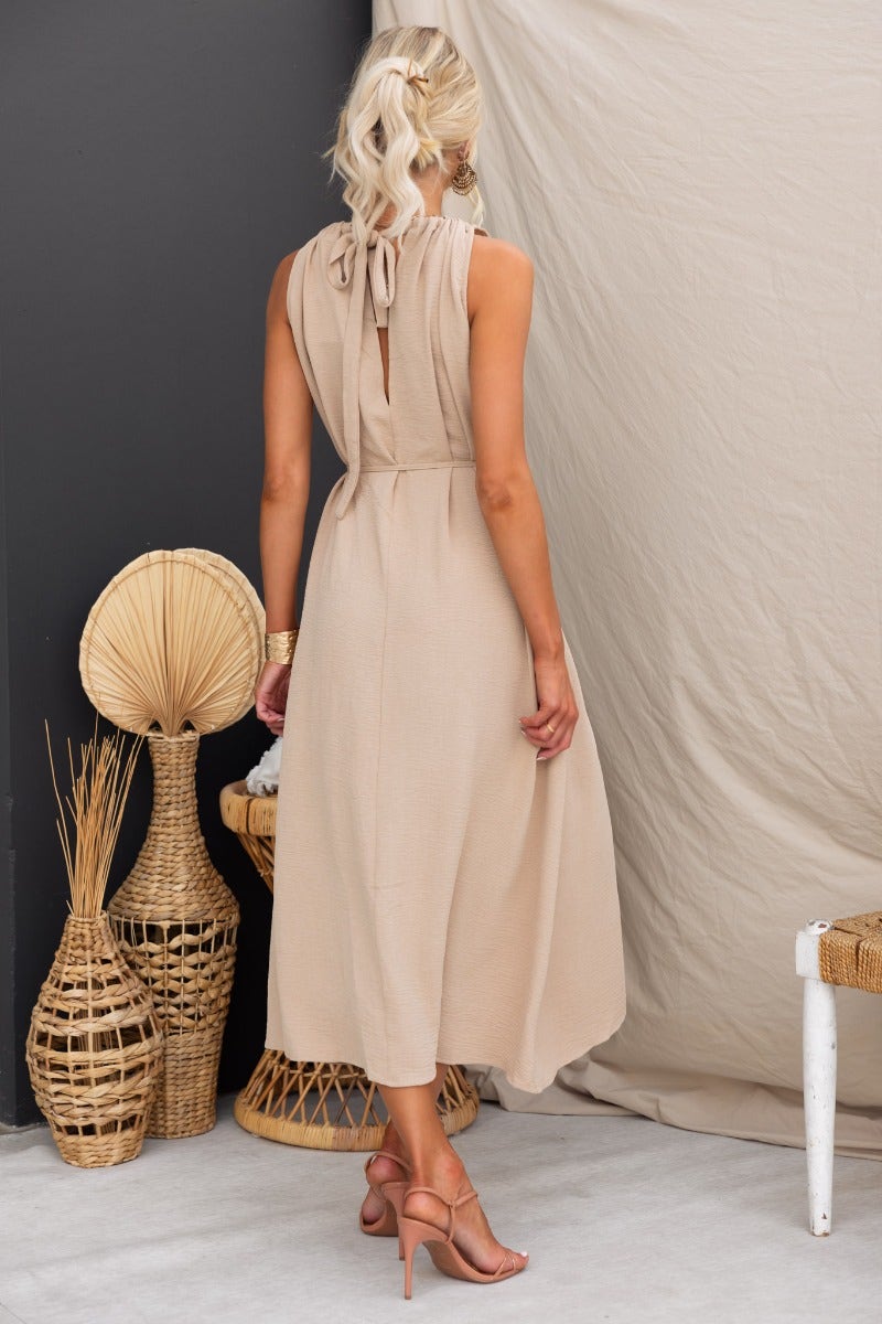 Back view of a woman in a beige Iwaki maxi dress with drawstring neckline and pockets, styled with strappy heels.