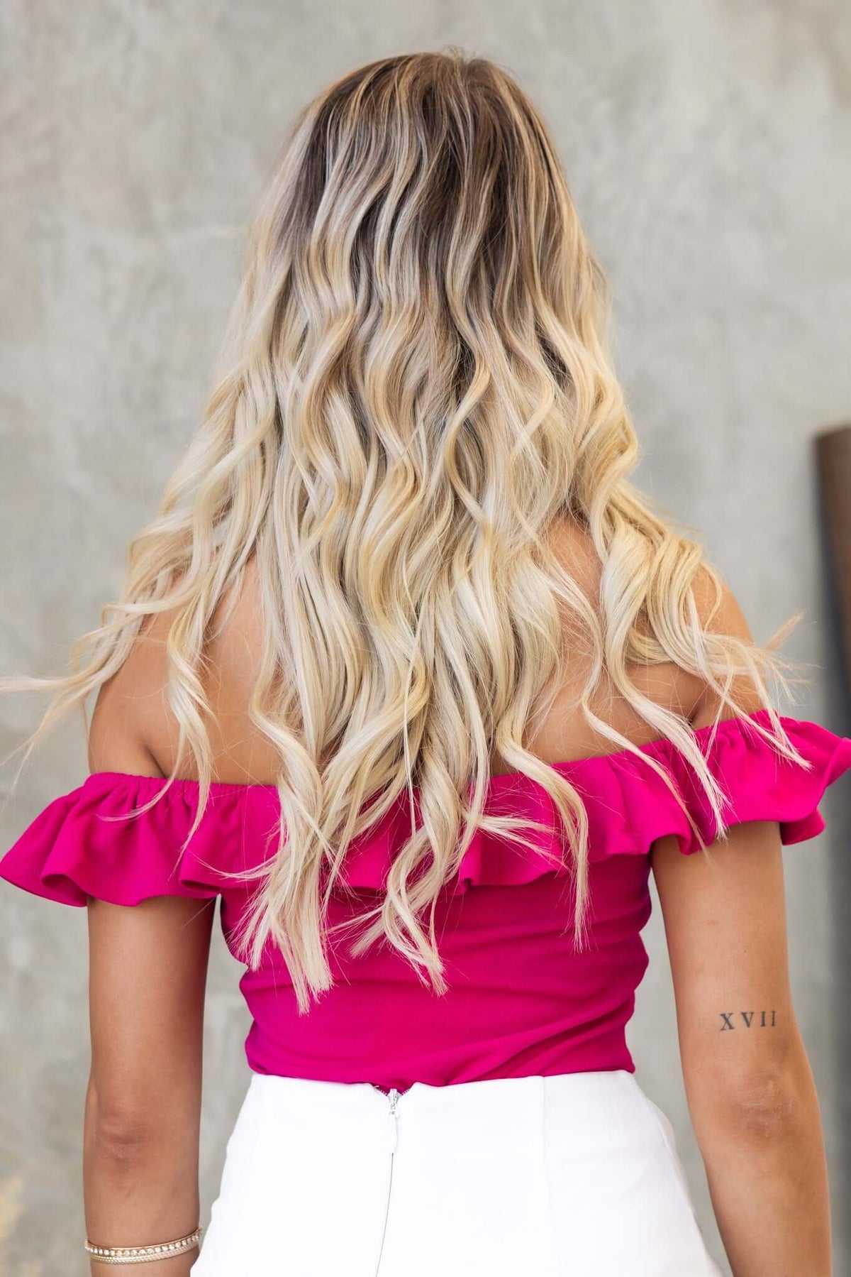 Back view of a model wearing a pink off-shoulder Bellar bodysuit with ruffle details and wavy blonde hair.