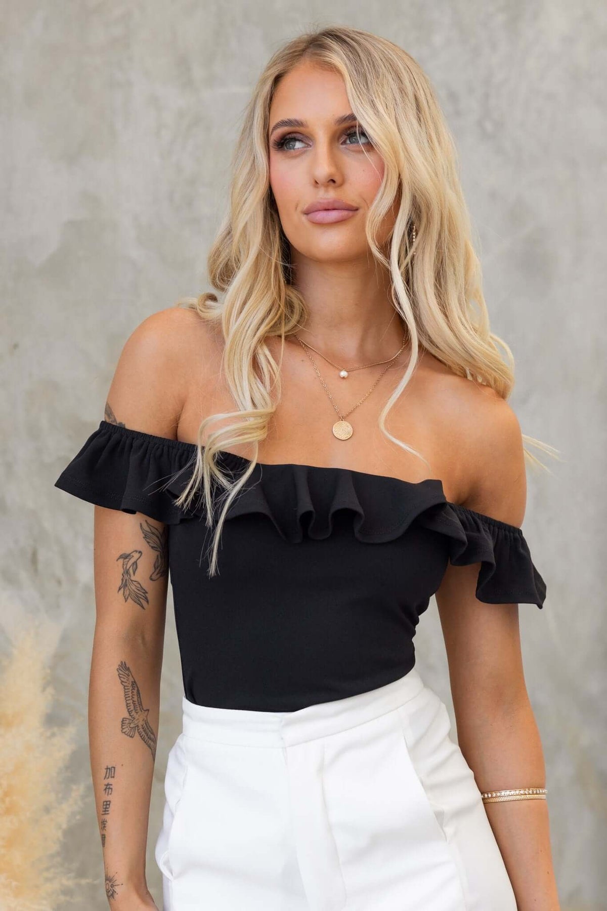 Model wearing a black off-shoulder Bellar bodysuit with ruffles and a stylish necklace, posing elegantly.